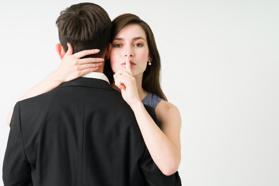 Woman with finger to lips making a silence gesture behind a man's back, both dressed formally