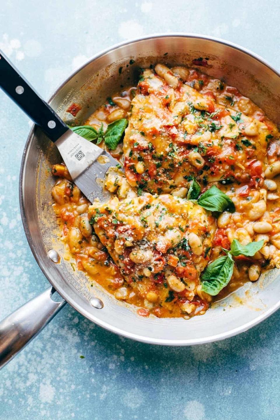 A skillet filled with fish in a creamy tomato-basil sauce with beans and a spatula