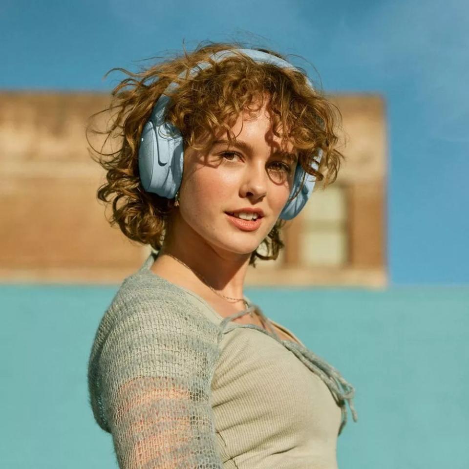 Person with curly hair wearing large headphones and a light knitted top smiles at the camera outdoors. Featured in an article on shopping for headphones