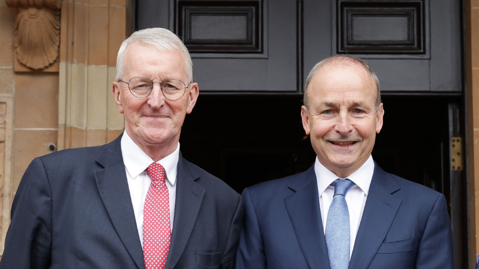 Hilary Benn in a red tie and white shirt with blue blazer met with Micheál Martin in blue blazer and white shirt blue tie, stood in front of doors of hillsborough castle 