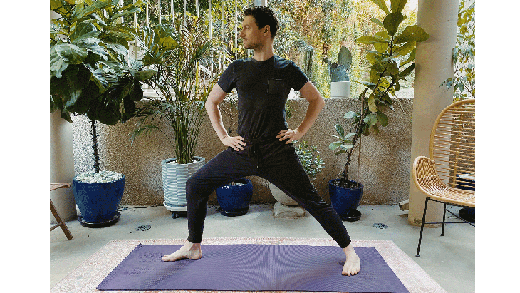 Man standing on a yoga mat with his hands on his hips in a variation of Warrior 2 Pose