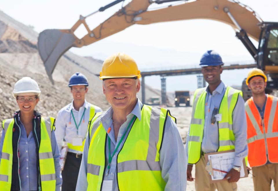 Construction workers standing in front of a backhoe. 