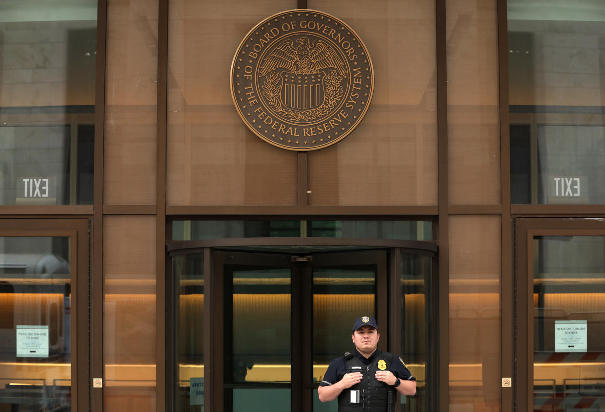 A guards stands at a building's entryway beneath a large seal reading: Board of Governors, the Federal Reserve System.
