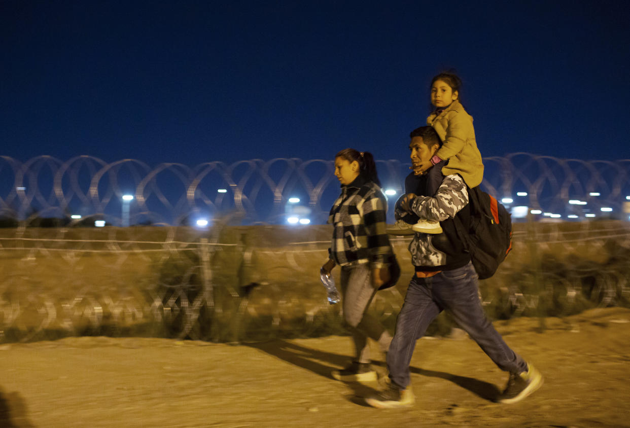 A migrant family from Peru, the man carrying a young girl on his shoulders, walk toward a gate in the border fence.