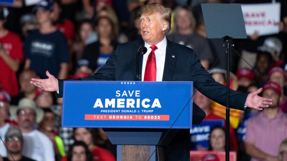 Donald Trump stands with arms outstretched behind a lectern that reads: Save America.