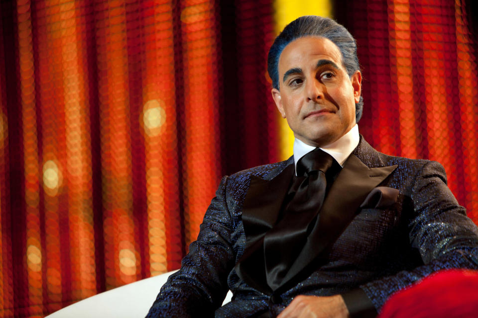 A man with slicked-back hair and wearing a formal, glittery jacket with a shiny black bow tie sits confidently against a vibrant, textured background