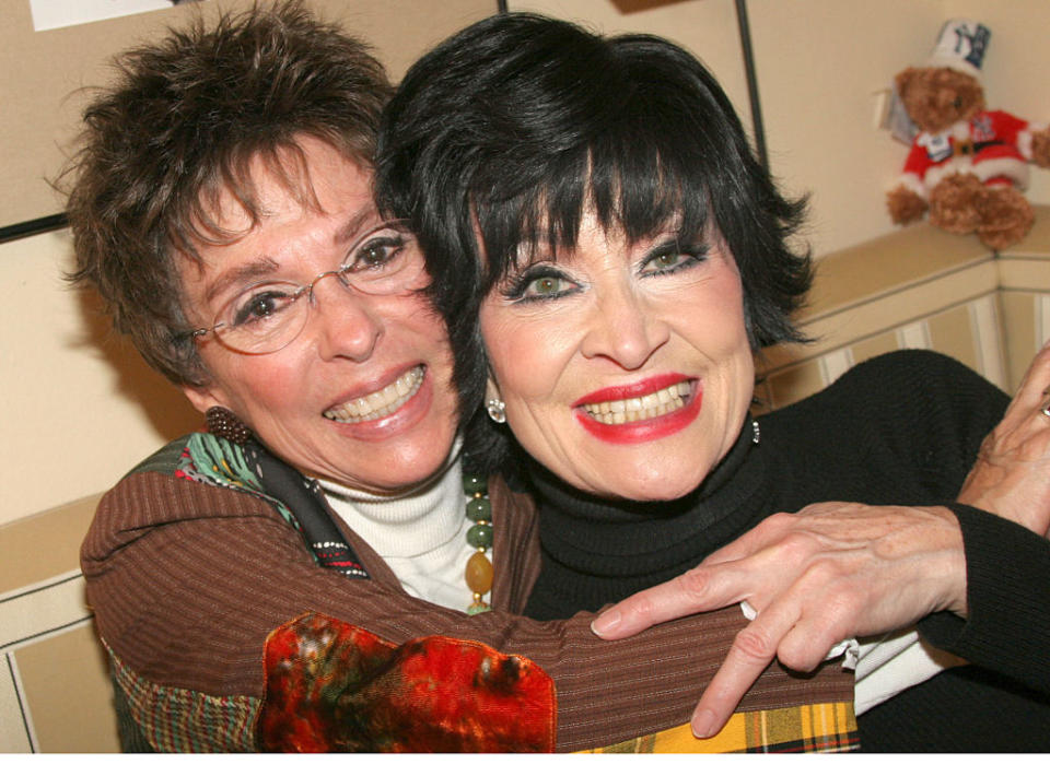 Two women embracing, smiling at the camera, one wearing a black top and the other a plaid jacket