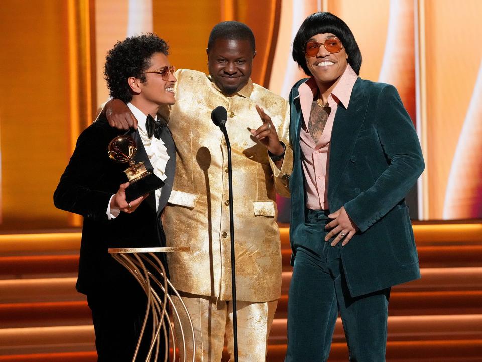 Bruno in a black tuxedo, Dernst in a gold jacket and pants, and Anderson in a dark green velvet suit, light pink wide collared button down, orange-tinted glasses, and a bowl cut wig.