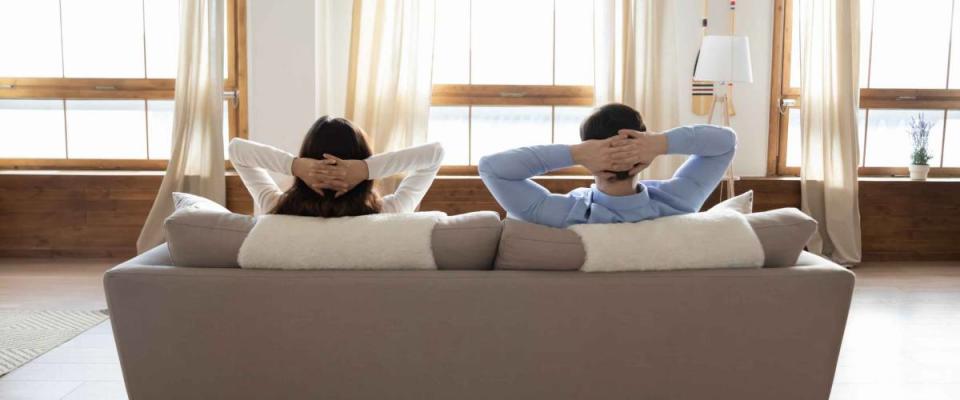 Rear view full length young couple relaxing on couch in modern home — not refinancing their mortgage.