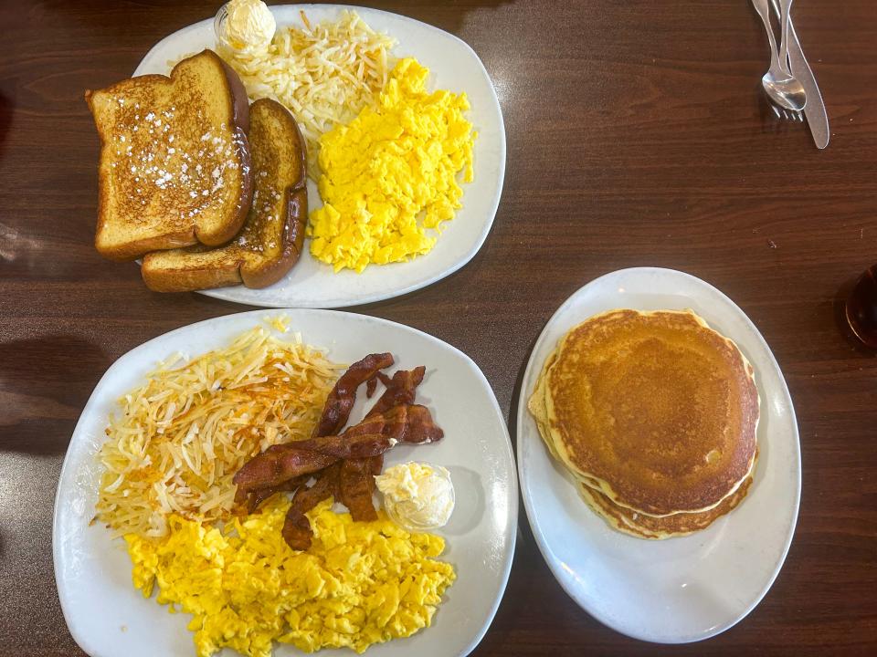 Three plates of breakfast food on a table at Perkins. Plates have French toast, hash browns, and scrambled eggs; hash browns, bacon, and scrambled eggs; pancakes