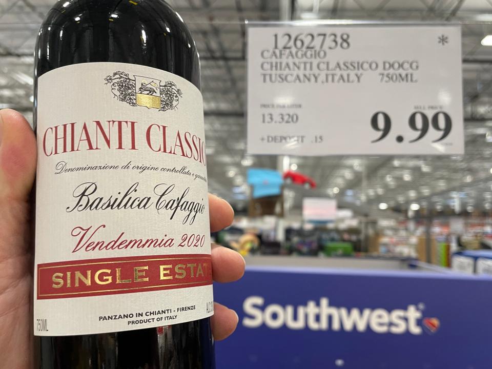 A hand holds a bottle of Chianti Classico wine in front of a price sign and Southwest display at Costco. The wine bottle is black and the label is cream