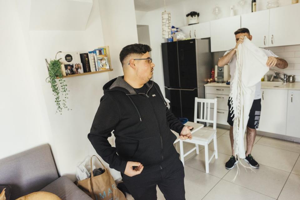 A man, left, in black clothing stands across from another man holding a shawl in an apartment