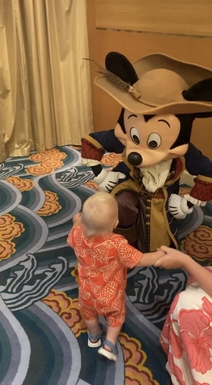 A baby in a patterned outfit approaches Mickey Mouse dressed in a pirate costume while holding an adult's hand