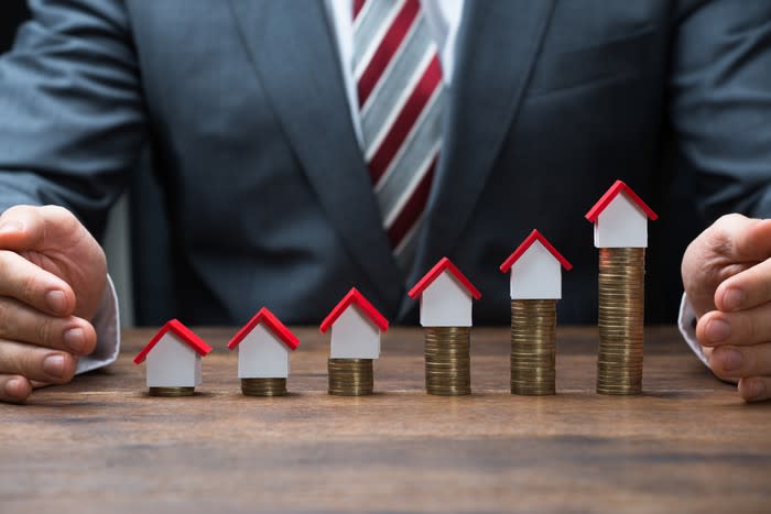 A person sitting behind toy houses on top of rising stacks of gold coins