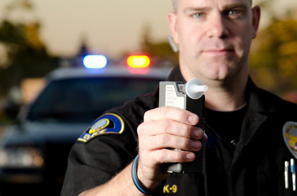 A police officer holding up a breathalyzer device, with his police vehicle in the background.