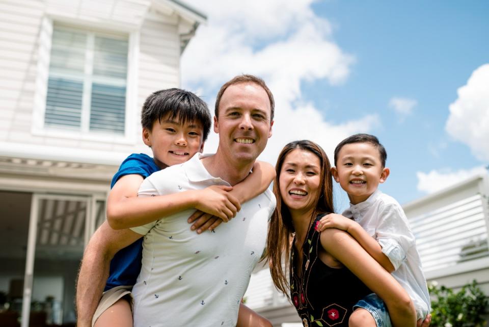 A mother and father, each with a son on their back, smile while standing in front of their house, ahead of the budget.