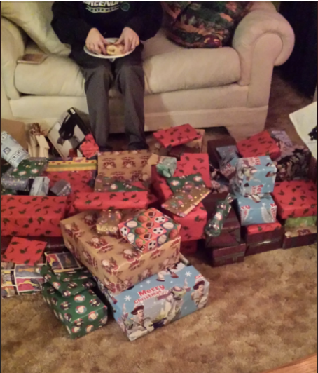 A child sits on a couch with a plate of food, surrounded by numerous wrapped presents, likely indicating a festive or holiday occasion