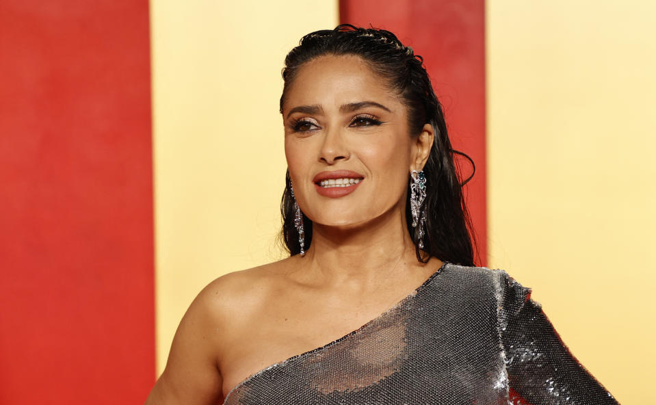 Salma Hayek at an event wearing a one-shoulder sparkling gown with dangling earrings