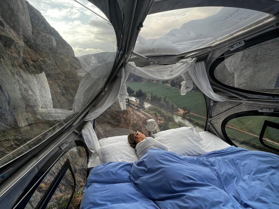 Victoria sleeps under a blue comforter in a hotel pod that hangs off the side of a cliff.
