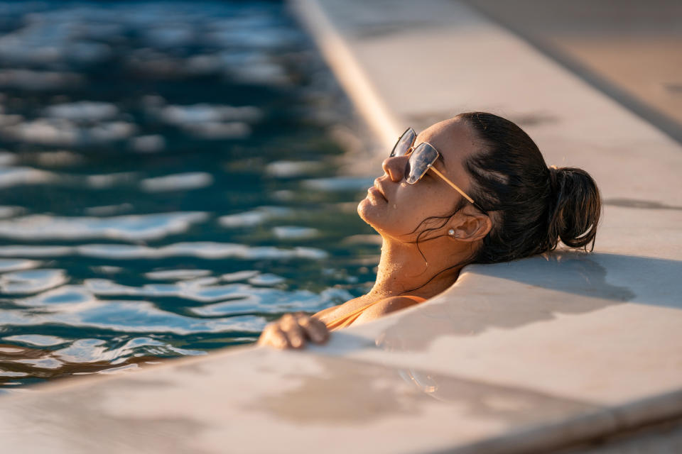 A woman wearing sunglasses is relaxing in a swimming pool, leaning against the edge with closed eyes and her hair tied up in a bun