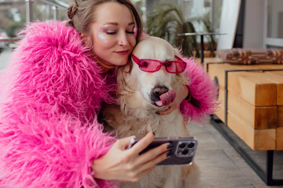 Woman in a pink fluffy fur coat takes a selfie with her dog wearing pink sunglasses.