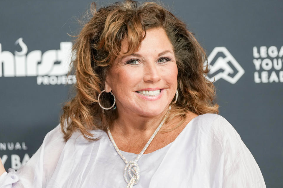 Celebrity Abby Lee Miller smiling at an event, wearing a white draped top with a large necklace and hoop earrings