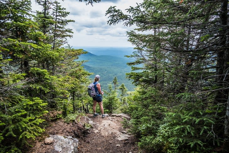 A woman is standing at an opening with a view point while climbing a mountain on the Appalachian Trail in Maine.