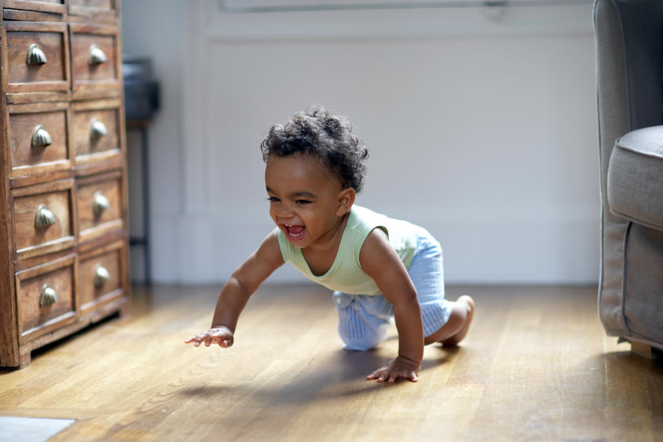 A joyful baby crawls on a wooden floor in a well-lit room, smiling and reaching forward