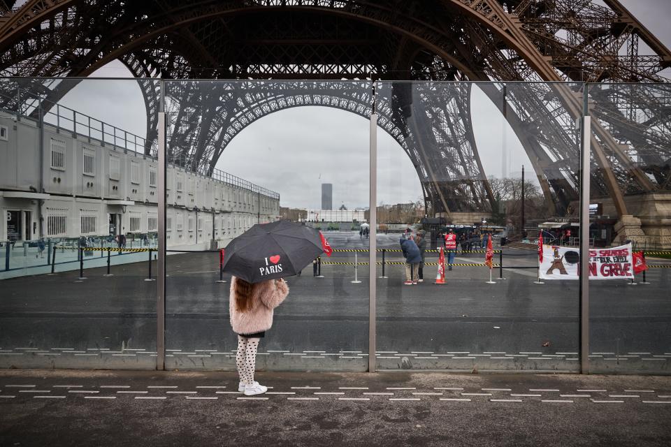 A pedestrian with an umbrella reading "I Love Paris" looks on next to the Eiffel Tower and a displayed banner reading "employees of the Eiffel Tower on strike" in Paris on February 19, 2024.