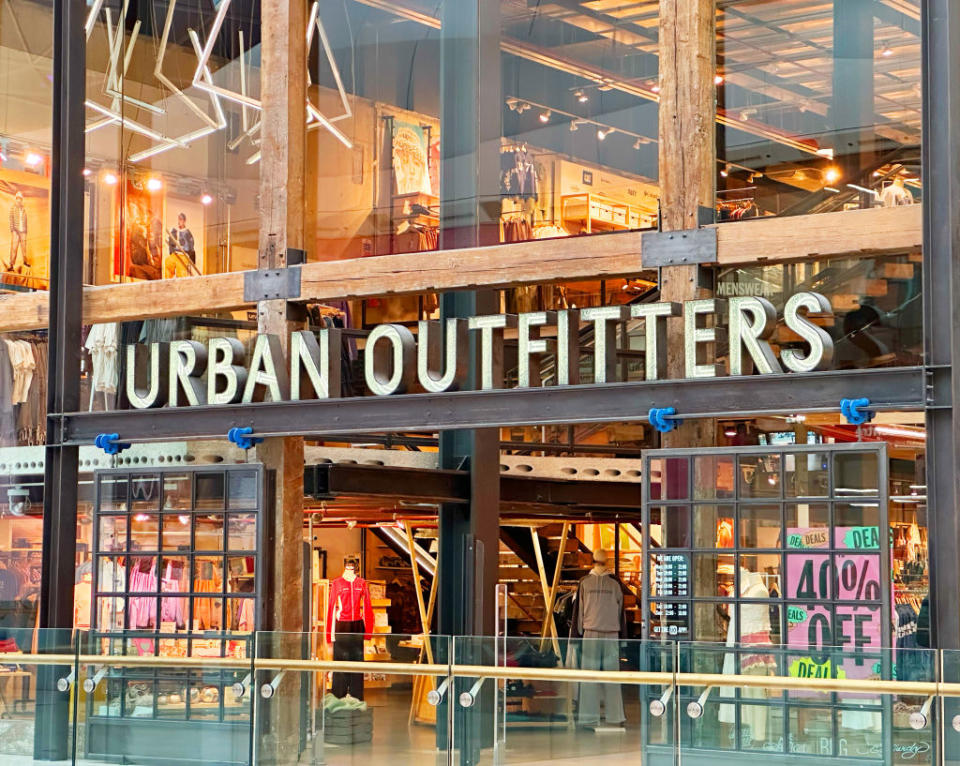 Exterior view of Urban Outfitters storefront, featuring display windows with clothing and interior decor items visible, and a sale sign in the window