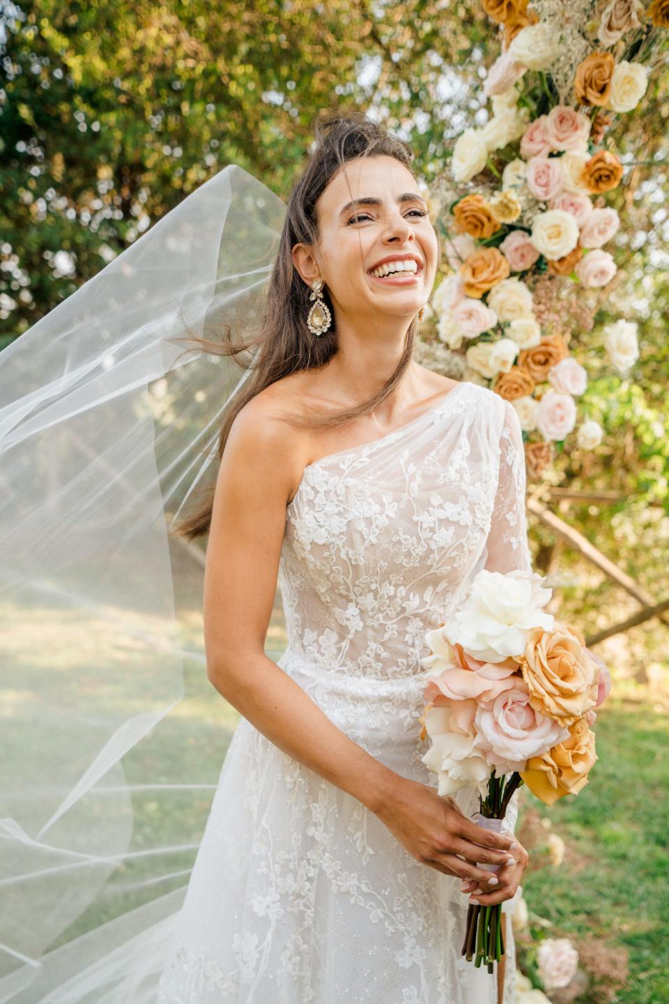 A bride smiles holding a bouquet of flowers as her veil flies in the wind.