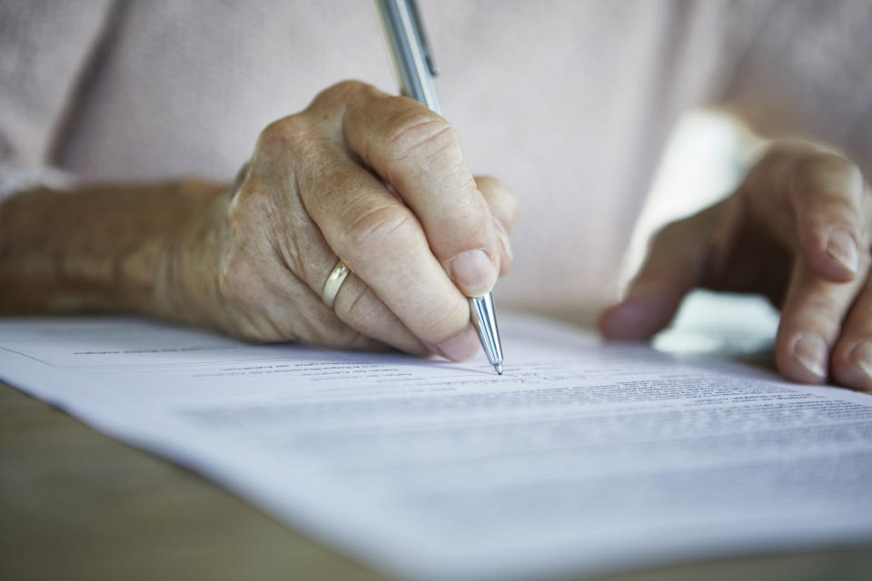 A close-up of an elderly person's hand holding a pen while signing legal documents. The person's hand displays a gold ring