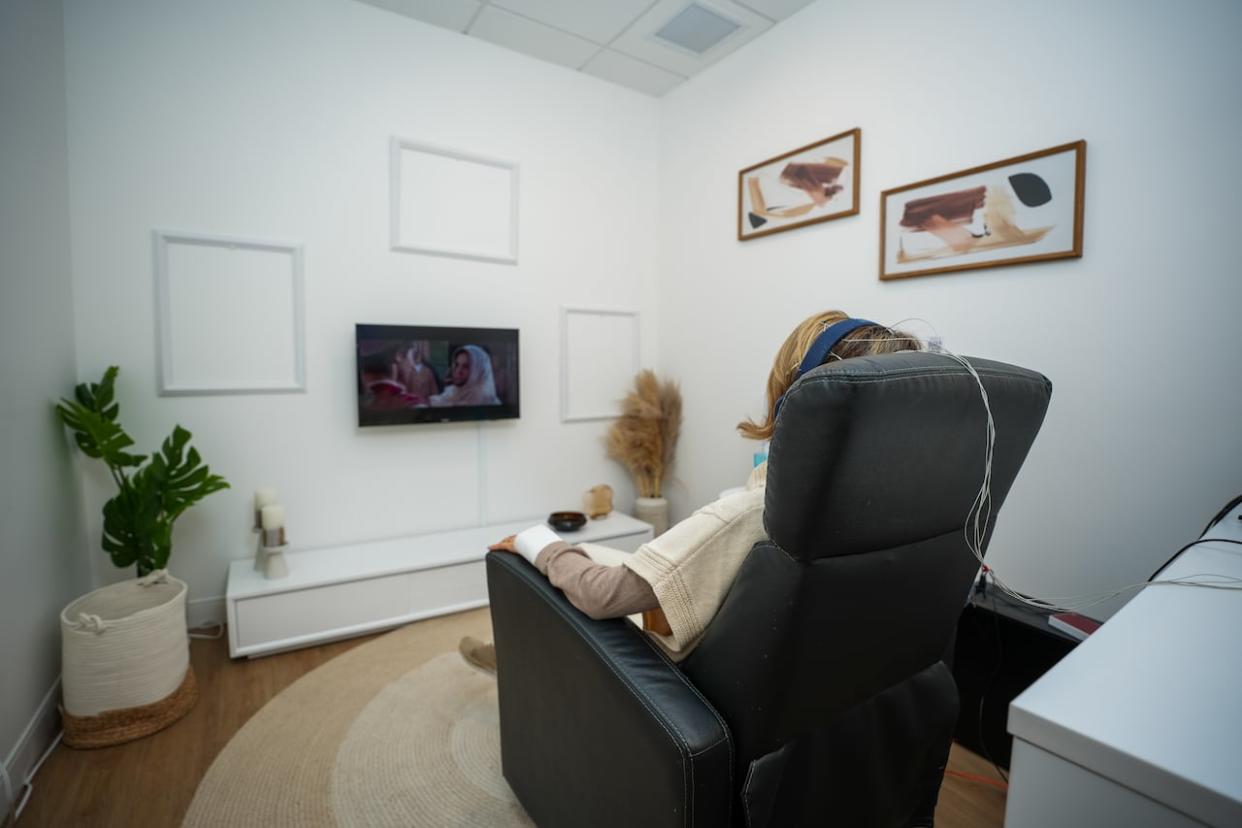 A woman demonstrates a technique used in neurofeedback therapy. Wires connect points of her head to an electroencephalogram (EEG) monitor on a computer, as she watches a film on a screen. (Submitted by Elumind Centres For Brain Excellence - image credit)