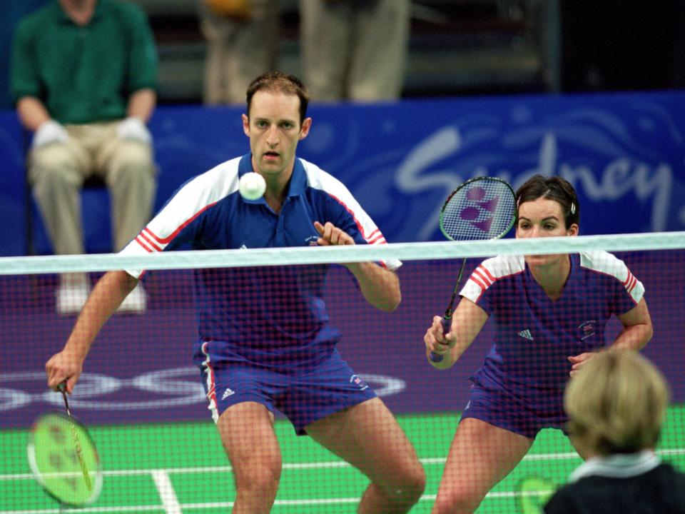 Badminton player gets in front of of the badminton from across the net while a player stands ready behind him.