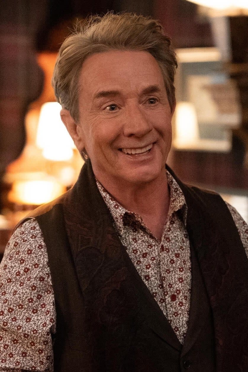 Martin Short stands smiling in a cozy, dimly-lit room with a piano and a pianist in the background. He wears a patterned shirt and dark vest