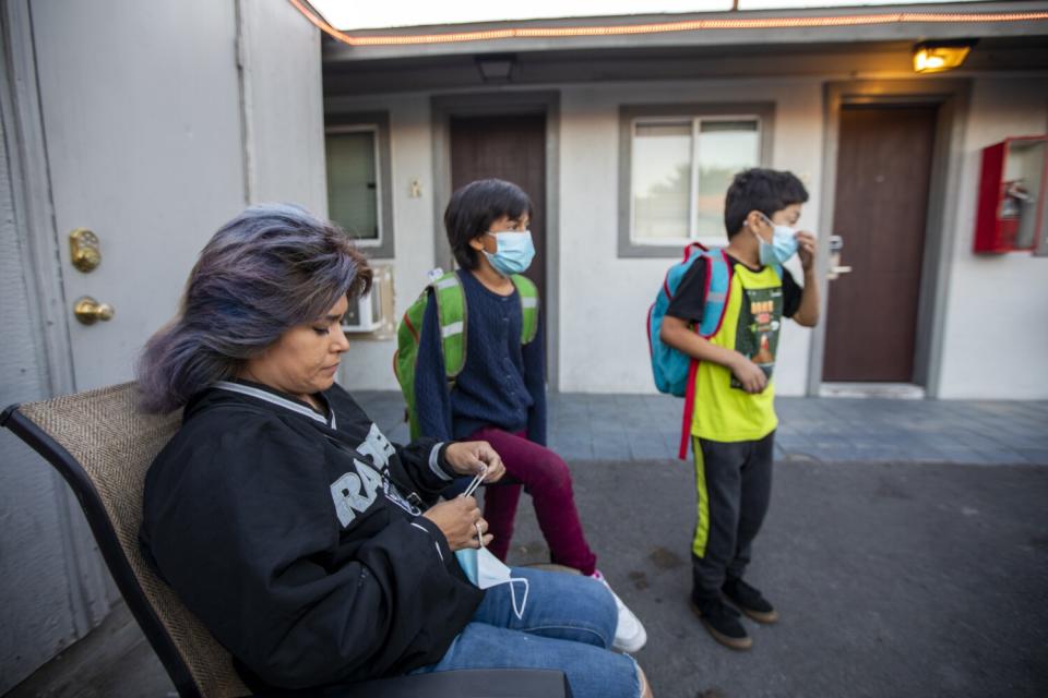 A woman sits in a chair while two young children walk past her with backpacks.