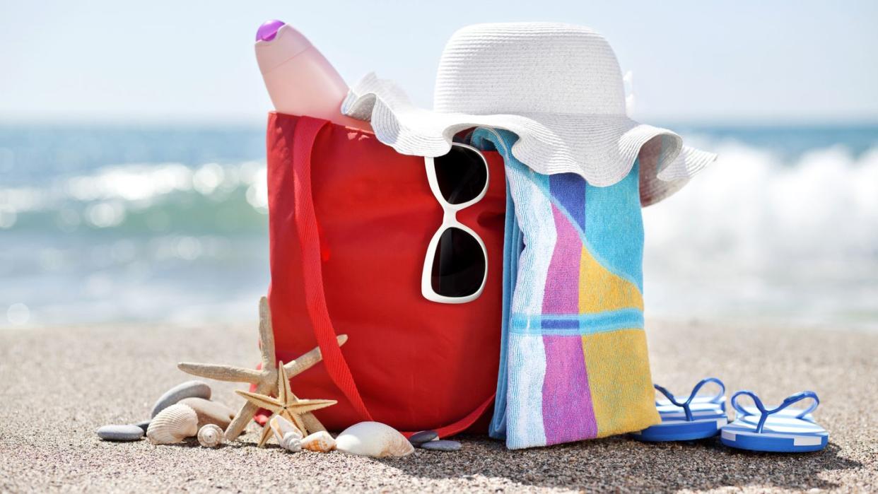 beach bag on the sand filled with sunglasses, a sunhat, sunscreen, and other objects