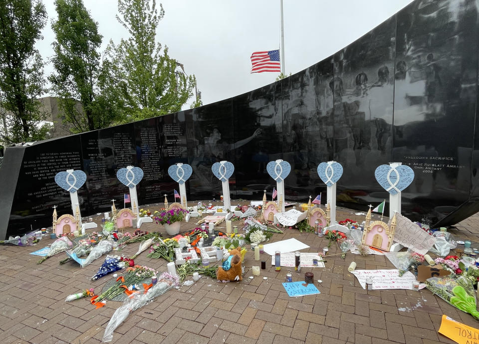 Flowers, candles and hand written signs lay on brick sidewalk alongside a black, curved memorial wall in front of an American flag at half-mast.