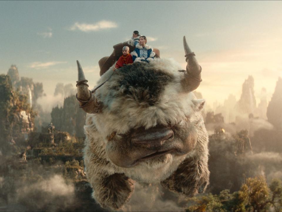 aang, katara, and sokka, teens clad in brightly colored clothing, sitting on top of appa, a large flying bison with fluffy hair, horns, and an arrow pattern on his forehead