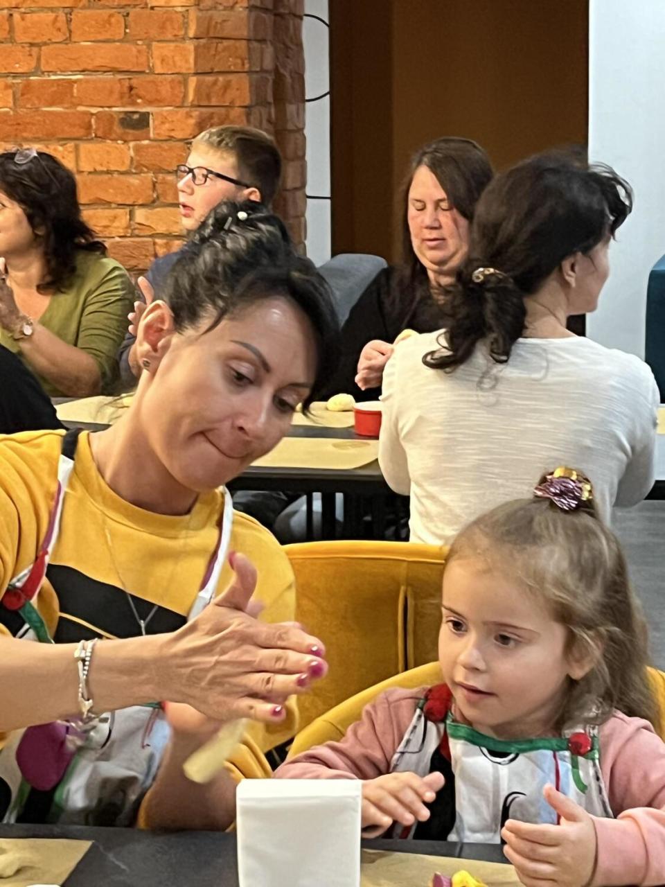 A woman with dark hair and a yellow top rolls dough as a young girl with light brown hair watches