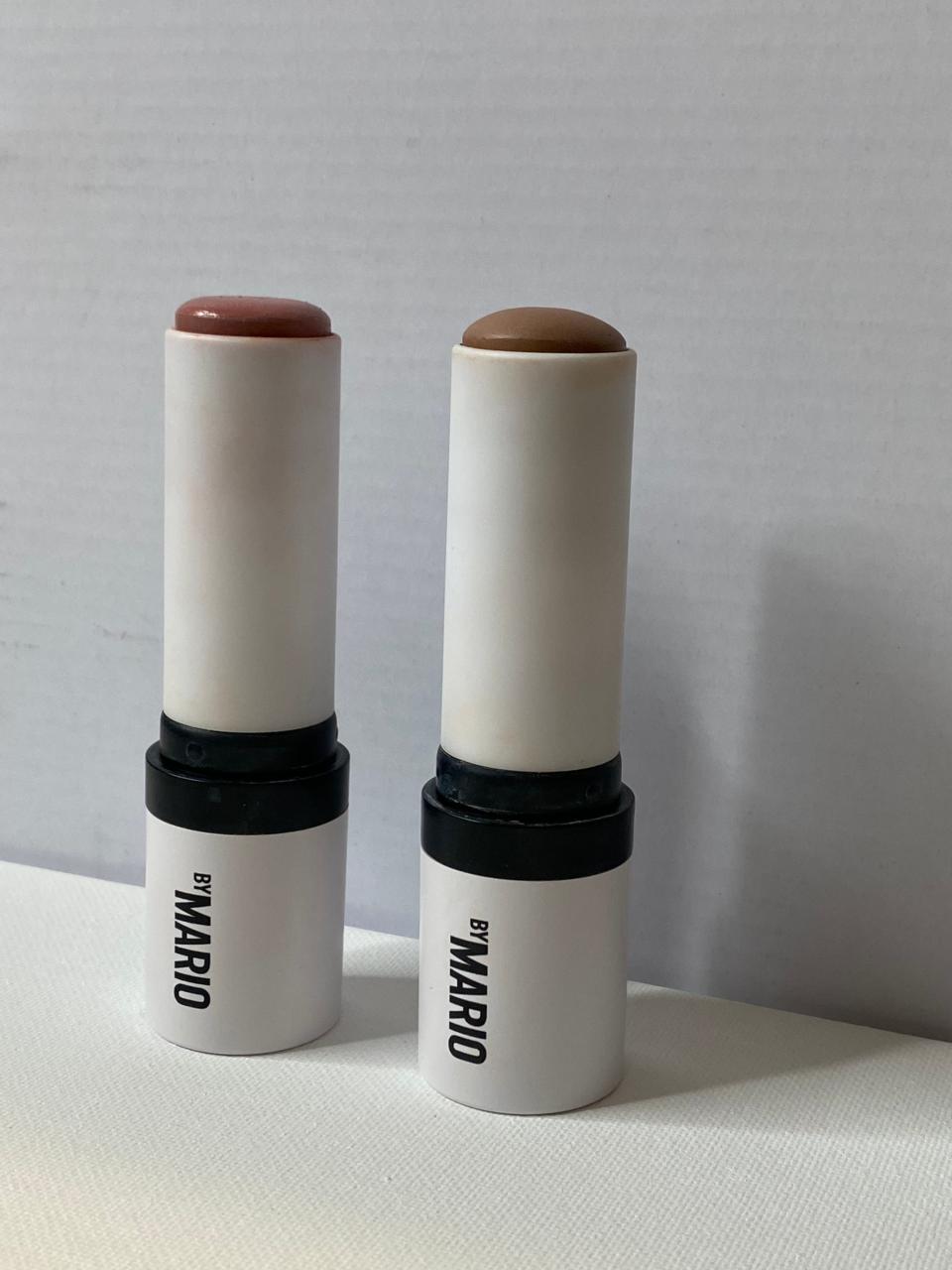 Two cream makeup sticks with "by MARIO" text on side. Product on the left is pink and the product on the right is brown.