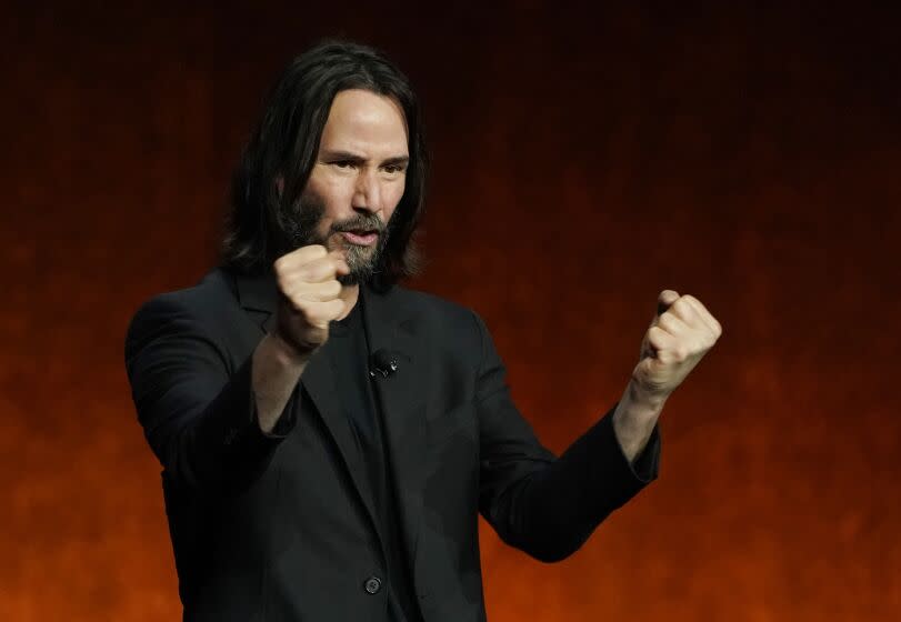 Man, Keanu Reeves, with long hair and bear, dressed in black blazer and shirt, clenching fists on stage