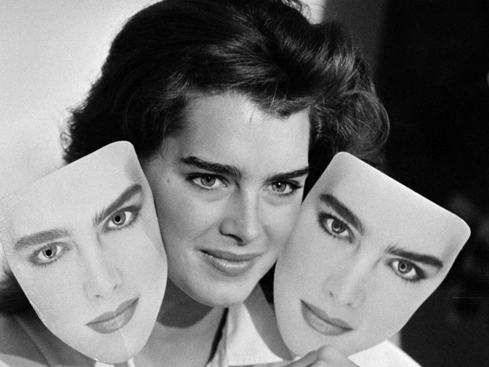 Brooke Shields holding up two masks of her own face at the end of a press conference in 1985