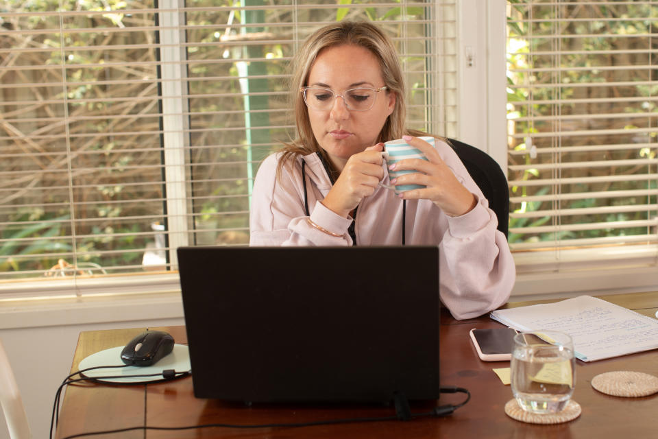 A women sits at the dining room table with a cup of coffee while working remotely on her laptop computer