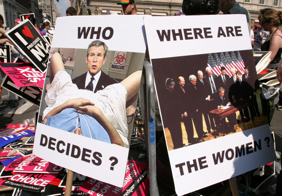 Two signs read "who decides?" and "where are the women?": the first showing George W Bush between the legs of a person on a medical examining table, the other showing a group of male politicians standing around Bush signing a document into law