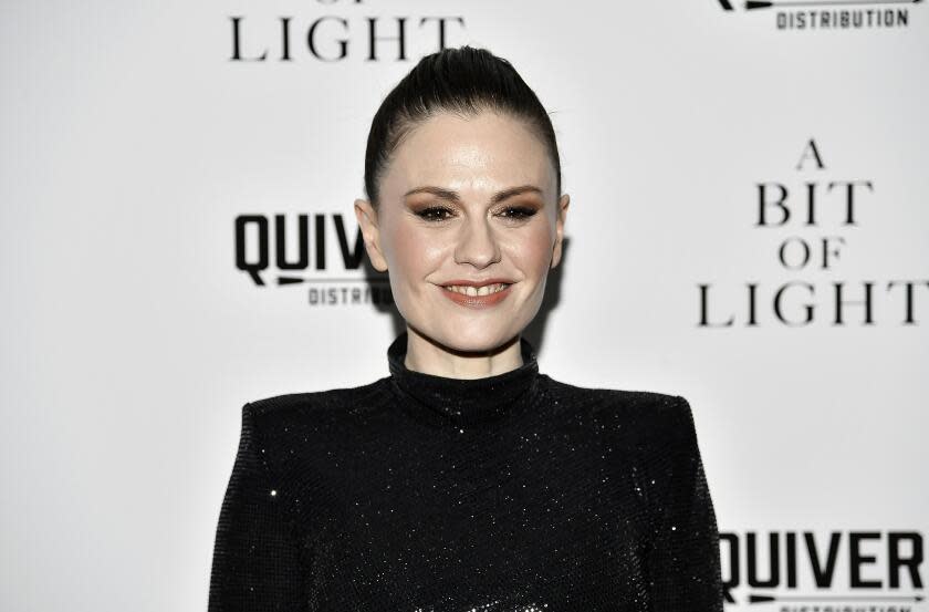 A woman with her dark hair slicked back wears a sparkly, black dress with shoulder pads. She poses against a white background