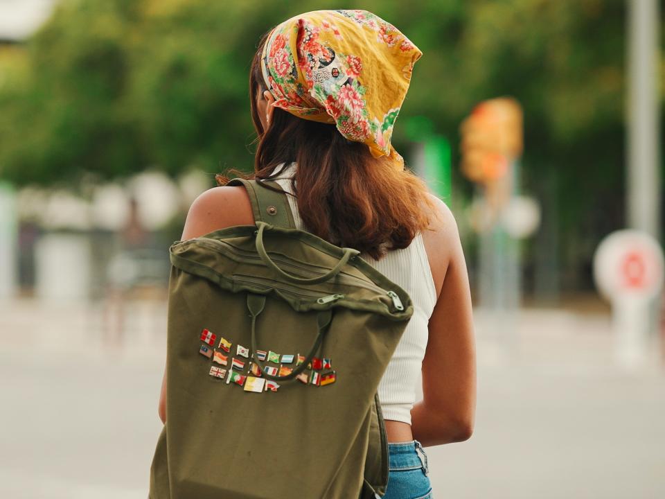 back of a woman walking down a street wearing a yellow bandana and carrying a green canvas backpack