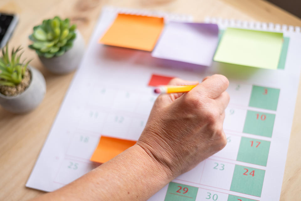 A hand writes on a calendar with colorful sticky notes at the top. Two small potted plants are on the table