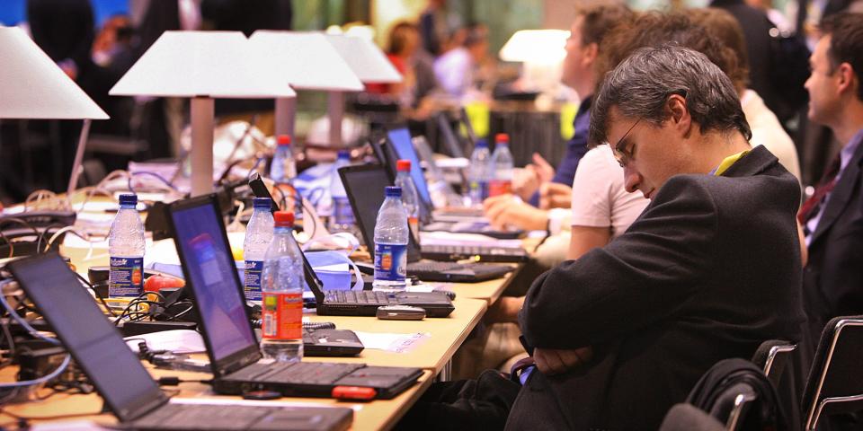 A mean wearing a suit and glasses asleep at a long desk where other people are using laptop computers.