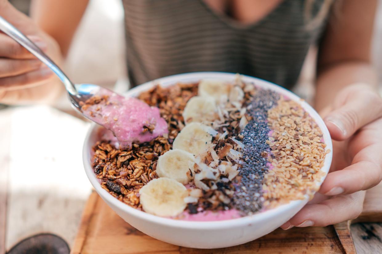 Closeup of quinoa breakfast bowl with a spoon in a white bowl, with a woman holding the spoon and bowl, with a blurred background of table and the torso of the woman